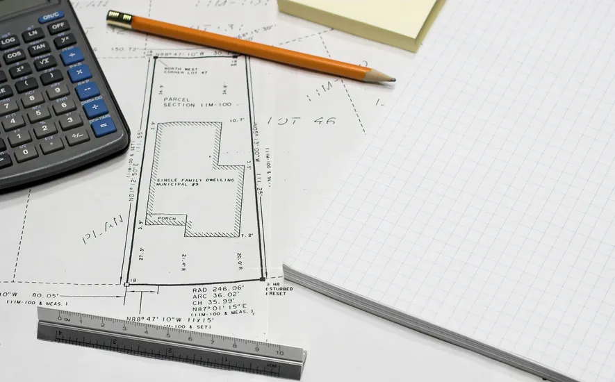 A calculator, architectural plans, a ruler, a pencil, sticky notes, and graph paper arranged on a workspace for project planning.
